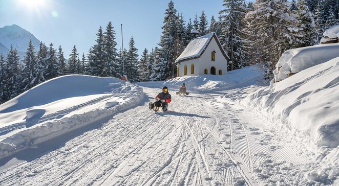 Rodler fährt die verschneite Rodelbahn Kappl an einer kleinen Kapelle vorbei